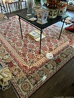 Full view of hooked area rug under black metal table, showing floral geometric pattern with red and cream dominant colors.