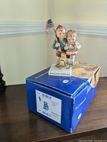 Porcelain Mj Hummel figurine of two children, one holding an American flag, sitting atop original box with "United States of America" inscribed on base.