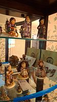 Glass shelf display with several Hummel figurines including the child on sled and others, showing packaging material nearby.
