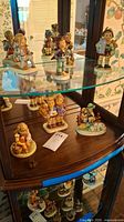 Display of seven Hummel porcelain figurines on glass and wooden shelves in a curio cabinet.