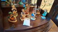 Eight Hummel figurines displayed on a wooden chair including children musicians and seated figures