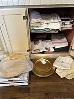 Photo of linen cabinet shelves showing stacks of folded table linens, mostly white and pastel colors, some with floral embroidery, along with gold-tone charger plates placed on the floor next to cabinet.
