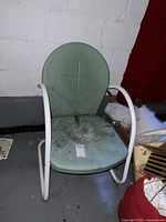 Green metal patio chair with white tubular metal frame shown against a basement floor and wall, visible weathering and dirt on seat and backrest.
