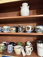 Shelf showing a variety of 21 ceramic mugs in various styles and colors, including floral, checkered, text, and images, plus a white ceramic vase on top shelf.