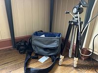 Photo showing Olympus E-520 camera, gray camera bag, and metal tripod arranged upright on floor corner.
