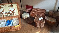 Wicker picnic basket open showing utensils, plates, cups, and napkins included, with three additional baskets and a jewelry box arranged around it.