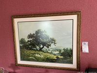 Photo showing framed landscape print with a tree and hills, set against a red wall.