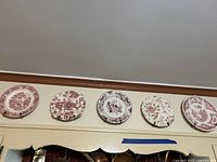 Five round plates showing distinct red and white vintage floral and cottage scenes on display above a cabinet.
