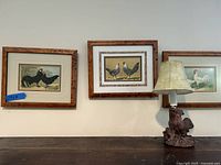 Three framed rooster prints and rooster lamp on a dark surface against a white wall.