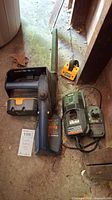 Photo showing the Ryobi chain saw, orange battery, and charger all placed on a concrete floor with visible use wear and dust.