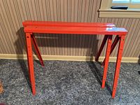 Two red metal saw horses against a wall on carpeted floor.