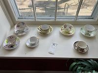 Seven teacups and saucers arranged on a windowsill showing various floral and gold designs