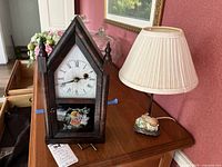 Front view of a vintage wooden mantel clock with pointed top and floral panel alongside a ceramic bunny lamp with beige shade