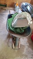 Blue metal wheelbarrow with wood handles and green water bag inside on a concrete floor