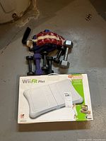 Wii Fit Plus balance board in original box shown with hand and wrist weights around it on a concrete floor.