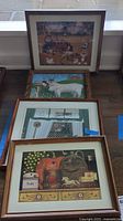 Four framed Charles Wysocki prints stacked leaning against a window, showing rustic Americana scenes including animals and kitchen items.