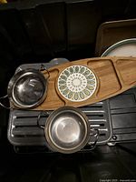 Small stainless steel pots, wooden serving tray with decorative inlays, and metal grilling tray inside bin