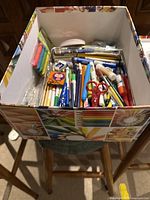 Top-down photo of a box filled with assorted art and craft supplies including pencils, pens, scissors, glue sticks, crayons, markers, and accessory packs.