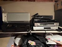 Photo showing stacked printers and electronic devices including DVD players and VCRs on a shelf.