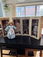Photo shows five porcelain dolls, four inside original boxes with storage wear, and one doll outside of box wearing a blue dress and white apron.