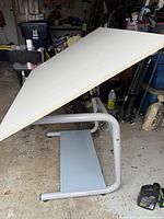 Large white tilt-adjustable drafting table on silver metal tubular frame base in garage/workspace.