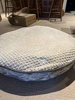 Photo of large round dog bed with light beige textured cover and visible zipper at side, showing Kirkland brand label, placed on carpeted floor.