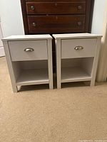 Pair of wooden off-white fiberboard side tables with drawers and open shelves, placed on carpeted floor, against a wall with a wooden dresser in the background.