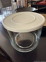 Pampered Chef glass trifle bowl with white lid over a counter surface, clear glass bowl cylindrical in shape, the lid embossed with Pampered Chef logo