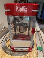 Front view of the Popflix popcorn machine featuring red top and base, clear housing, and visible metal kettle inside.