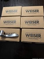 Six Weiser brand door handles each on top of individual branded cardboard boxes.