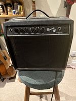 Front view of the Squier 15 amplifier showing control panel with knobs and inputs, black grille with logo, and top handle.