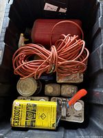 View inside heavy-duty black plastic storage bin showing red plastic gas can, coiled orange extension cord, multiple cans of liquids including Creosote wood preservative liquid, and various small containers.