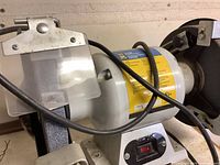 Side view of bench grinder showing grinding wheel, safety shield, and power cord