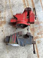 Photo of two small metal bench vices placed on a surface, one red and one blue, showing signs of wear and use.