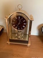 Front view of brass shelf clock showing brown Seiko dial with white Roman numerals, brass case with arched top, and four-ball pendulum inside glass panels.