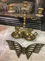 Photo showing one three-arm brass candlestick holder with white accent disks underneath small candle cups, two smaller single candle holders with scalloped edge trays, and a cut-out brass butterfly decoration.