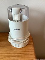 Front view of white Salton electric coffee grinder with clear plastic lid and power cord coiled around base, placed on wooden surface.