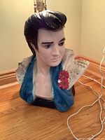 Front view of ceramic Elvis bust lamp showing the bust form, painted facial details, blue jacket, and electrical cord.