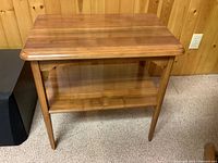 Clear photo showing the front and side of a small brown wooden occasional table with two tiers and tapered legs against a wood-paneled wall.