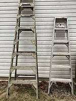 Two aluminum step ladders standing upright against a siding wall, showing paint marks and surface corrosion.
