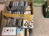Box of numerous CDs with visible album covers including a DVD or CD with photo of three people and title 'Love Through the Decades'. Appears to be a mixed music collection.