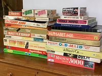 Photo showing stacked vintage board games including Rack-O, Yum, Perquackey, Tiddly Winks, Rummikub, Raggedy Ann, and others.