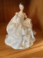 Front angle of Royal Doulton porcelain figurine showing seated woman in white dress with gold floral details, holding part of her dress.