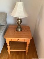 Oak side table next to a couch showing drawer knob, lower shelf, and top surface with lamp (lamp not included)