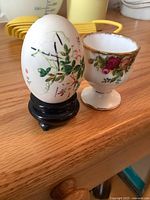 Decorative painted egg on black wooden stand beside a ceramic floral egg holder with gold trim, showing details of design and condition.