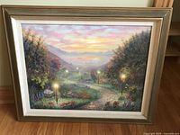 Framed oil painting showing a pathway lit by glowing lamp posts during sunset with detailed landscape including trees, flowers, and mountains.