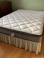 Queen mattress with quilted white cover, raised pattern stitching, thick dark trim edges, placed on boxspring with white skirt