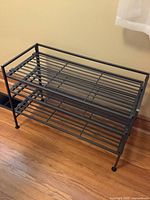 Black metal shoe rack with multiple slatted shelves shown against a tan wall on wooden floor.