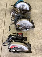 Three corded circular saws stacked together on the floor showing Makita, Craftsman, and Skil brands with cords tied.