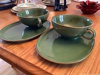 Two matching green ceramic soup plate sets each with bowl and oval underplate on a wooden table.